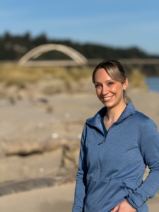 Medical Care Team Member Michaela on an southern Oregon beach with sand, ocean, and bridge to Bandon, Oregon. Blue sky is visible.