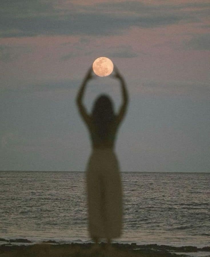 Silhouette of a person holding up the moon against a serene ocean backdrop during twilight.